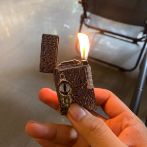 Bật lửa Devil’s Eye tông đen cầm trên tay, mở nắp và đang cháy với ngọn lửa đôi