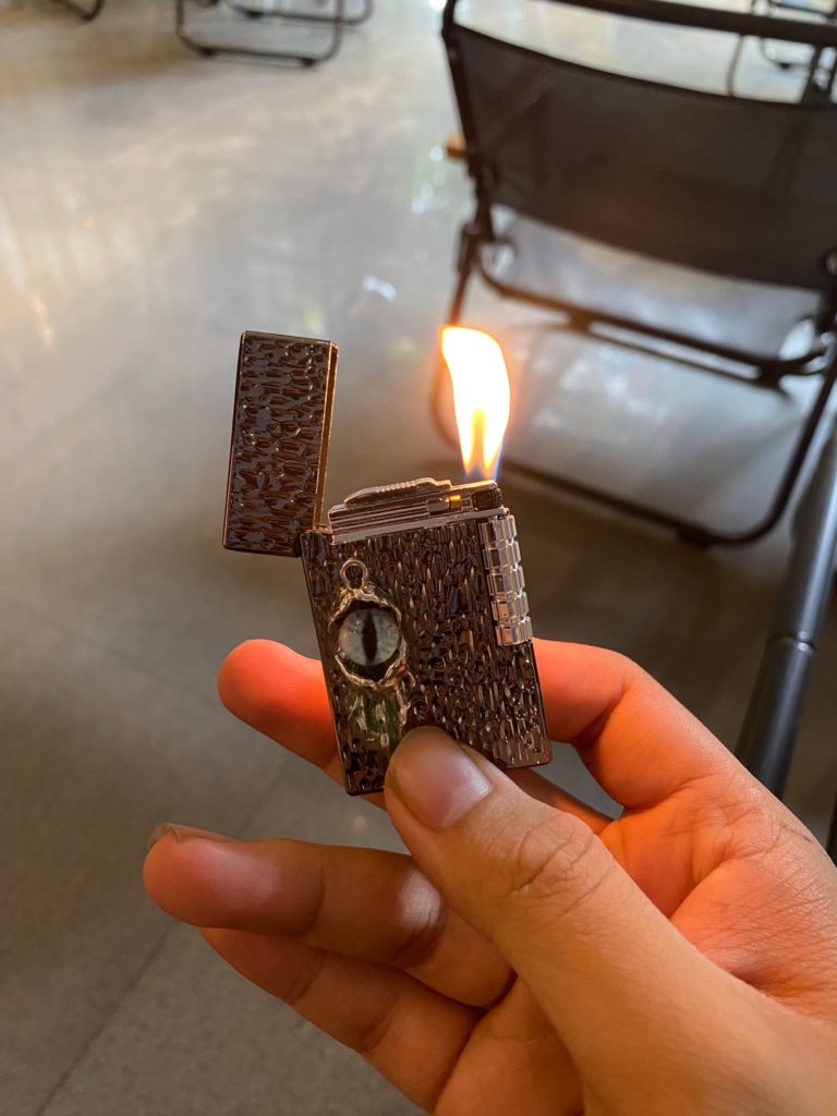 Bật lửa Devil’s Eye tông đen cầm trên tay, mở nắp và đang cháy với ngọn lửa đôi
